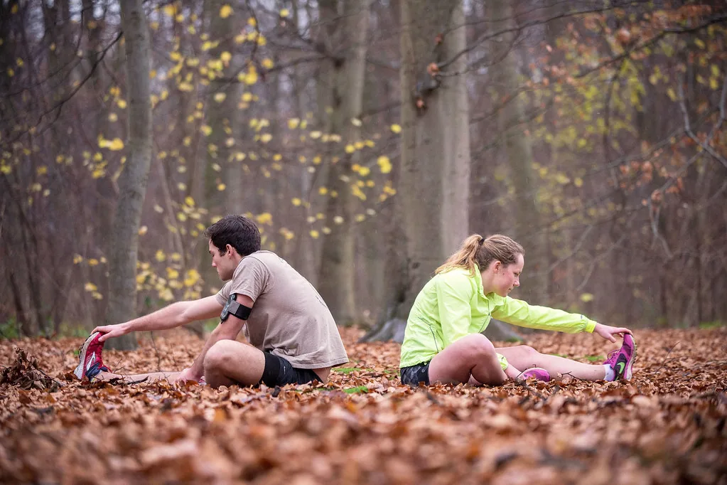 Kaksi juoksijaa venyttelee syksyisessä lehtimetsässä